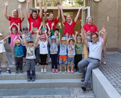 Kita-Mitarbeiterinnen und Grimmas Oberbürgermeister Tino Kießig mit Kindern der Einrichtung vor dem Eingang halten die Hände nach oben